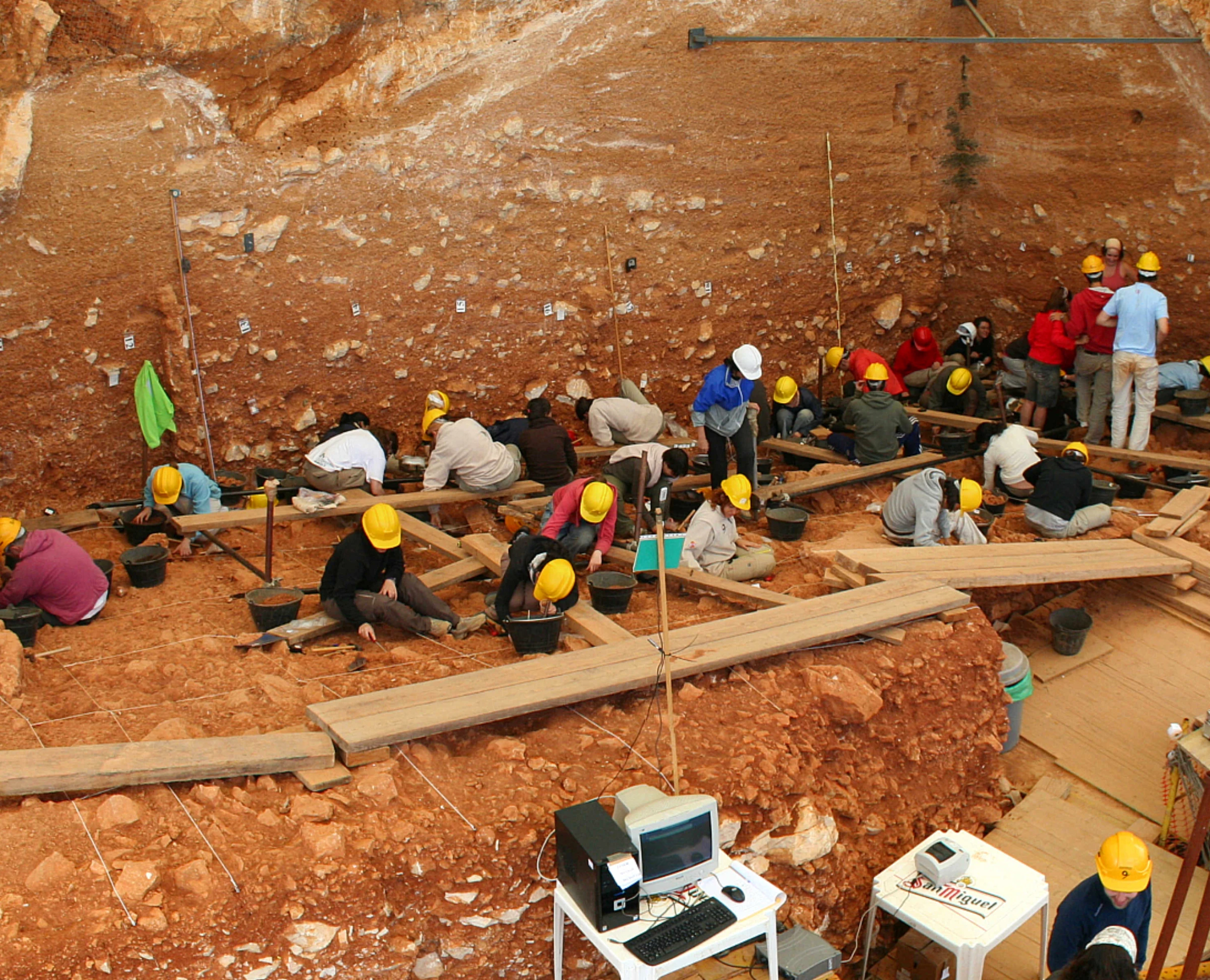 Archaeological excavation site showing layers of earth and artifacts being carefully uncovered with scientific instruments and documentation tools